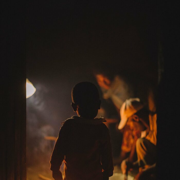 a young boy silhouetted in front of a fire on the ground outside of his home