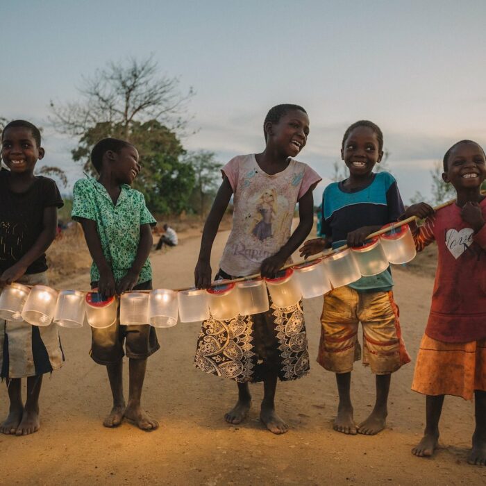 laughing and smiling young kids stand barefoot on a dirt road and hold a stick with 14 solar lanterns that they just received