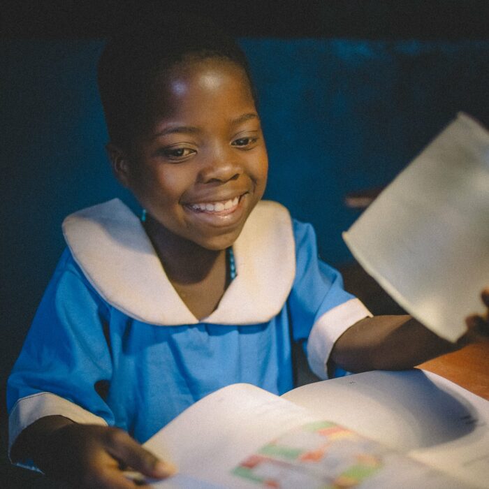 a young girl in a blue and white dress smiles while reading with lights in her home that were just donated by IAPW