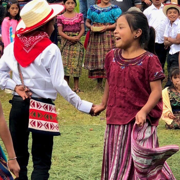 two young people dance in brightly colored traditional Guatemalan outfits on grass, surrounded by smiling and singing kids in similar outfits