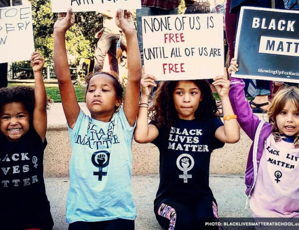 a diverse group of young girls hold sings above their heads with protest signs. one of which reads "none of us is free until all of us are free", and "Black Lives Matter"