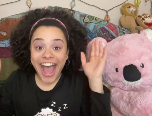 a young girl raises her hands in the air while laughing with a pink stuffed koala bear in bed next to her