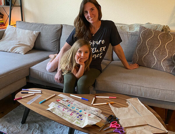 a young mother with a pretty smile sits with her cute daughter on a couch behind a modern coffee table covered in an art project and pens and small scissors that the young girl is working on