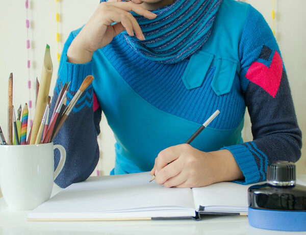 photo of a woman from the neck down to the desk in a modern blue sweater writing a letter. With a cup full of pencils and paint brushes, an ink well, and a book that she is writing in