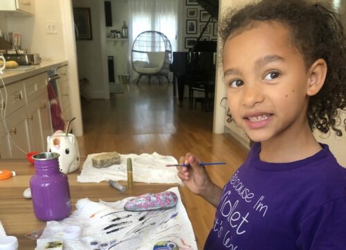 a young girl in a purple tee shirt smiles at the camera while painting rocks with faces in her kitchen