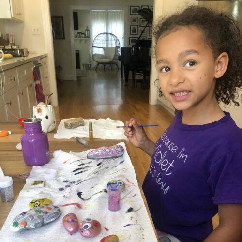 a young girl in a purple tee shirt smiles at the camera while painting rocks with faces in her kitchen