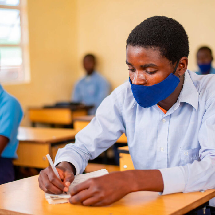 Mbongozi Student with a mask on sitting at his desk in school while writing