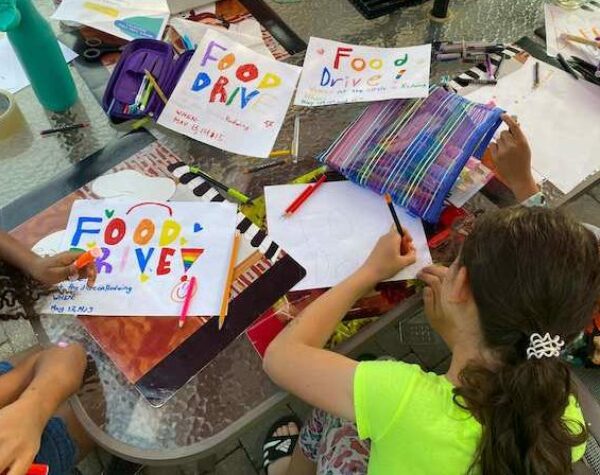 three young girls sitting on a patio with a table covered in flyers and pens for their food drive