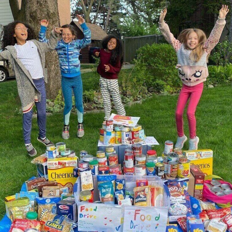 a diverse group of young kids jump in celebration behind a pile of food they collected to donate
