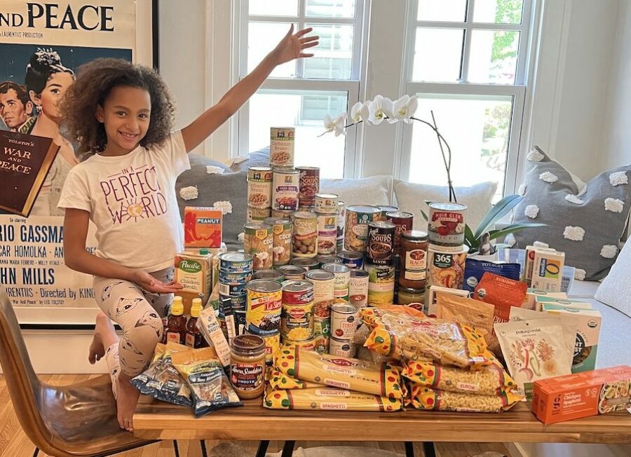 a young girl smiles and poses behind a table covered in food that she collected to donate