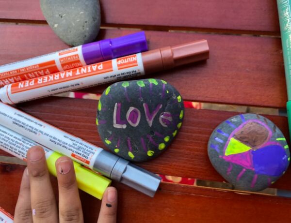 childrens hand with paint on the fingers and paint pens on a table next to rocks that have been decorated in bright colors with designs and hearts and the word Love
