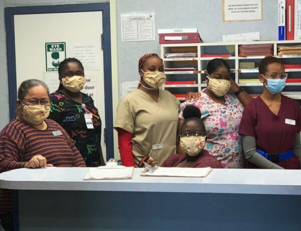 a group of nurses with masks on pose with a young girl behind a desk with masks that were donated