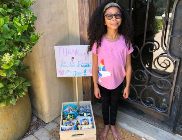 a young girl smiling with a box of donated items she collected standing in front of a door